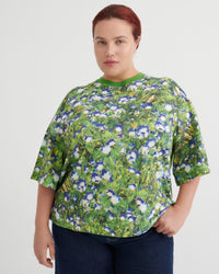 A woman wearing a short-sleeve crewneck top with a green, blue, and white floral print, paired with dark blue jeans, against a plain light gray background.