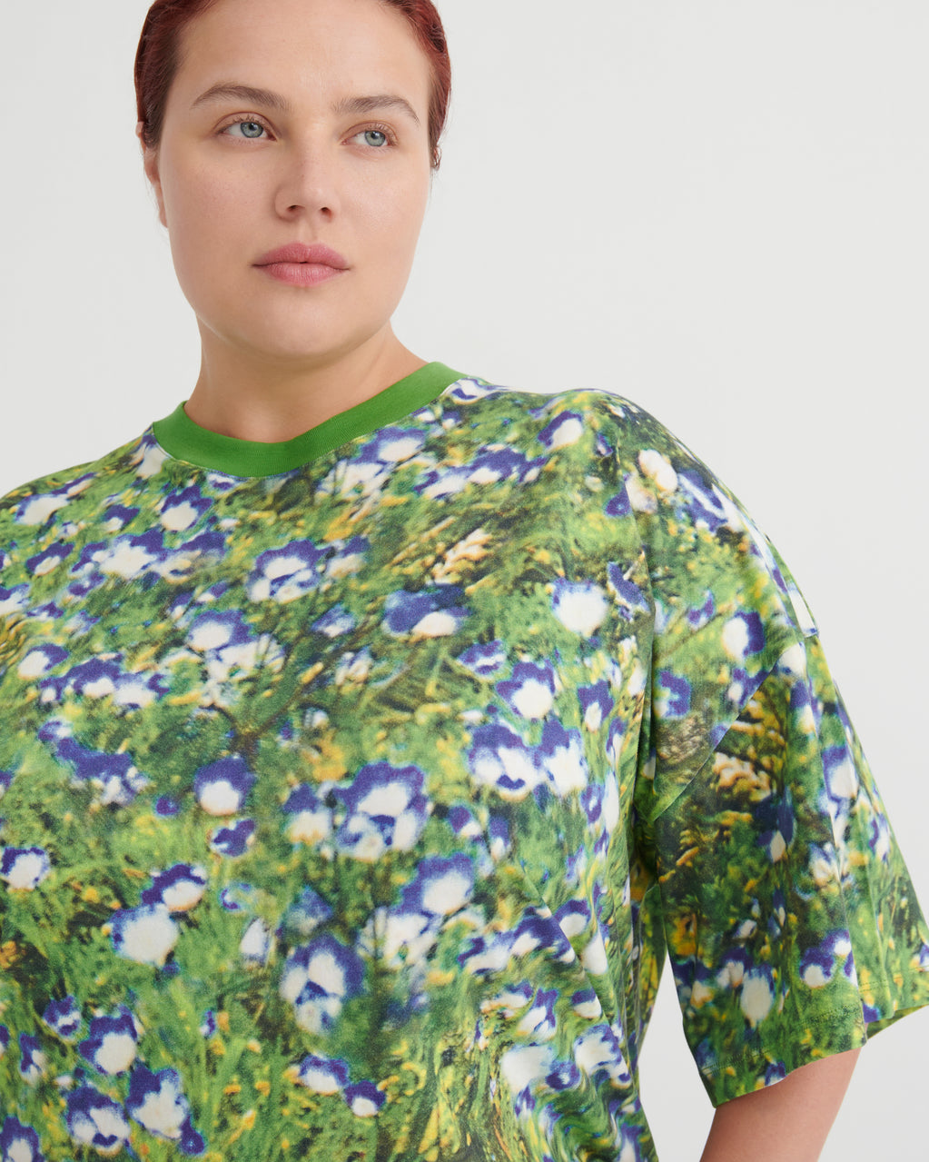 A close-up of a model wearing a short-sleeve crewneck t-shirt with an all-over floral print inspired by Van Gogh's flowers, featuring green, blue, and white blossoms on a textured green background.
