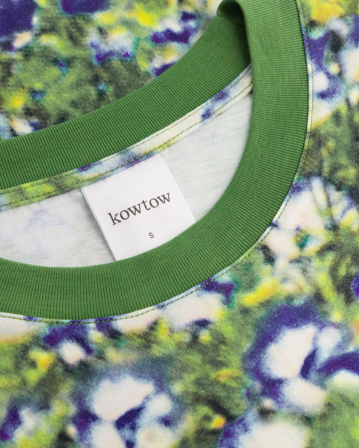 Close-up of a ringer t-shirt neck showing a green ribbed collar and a white brand label that reads 'kowtow'. The shirt features a vibrant green, purple, and yellow tie-dye pattern.
