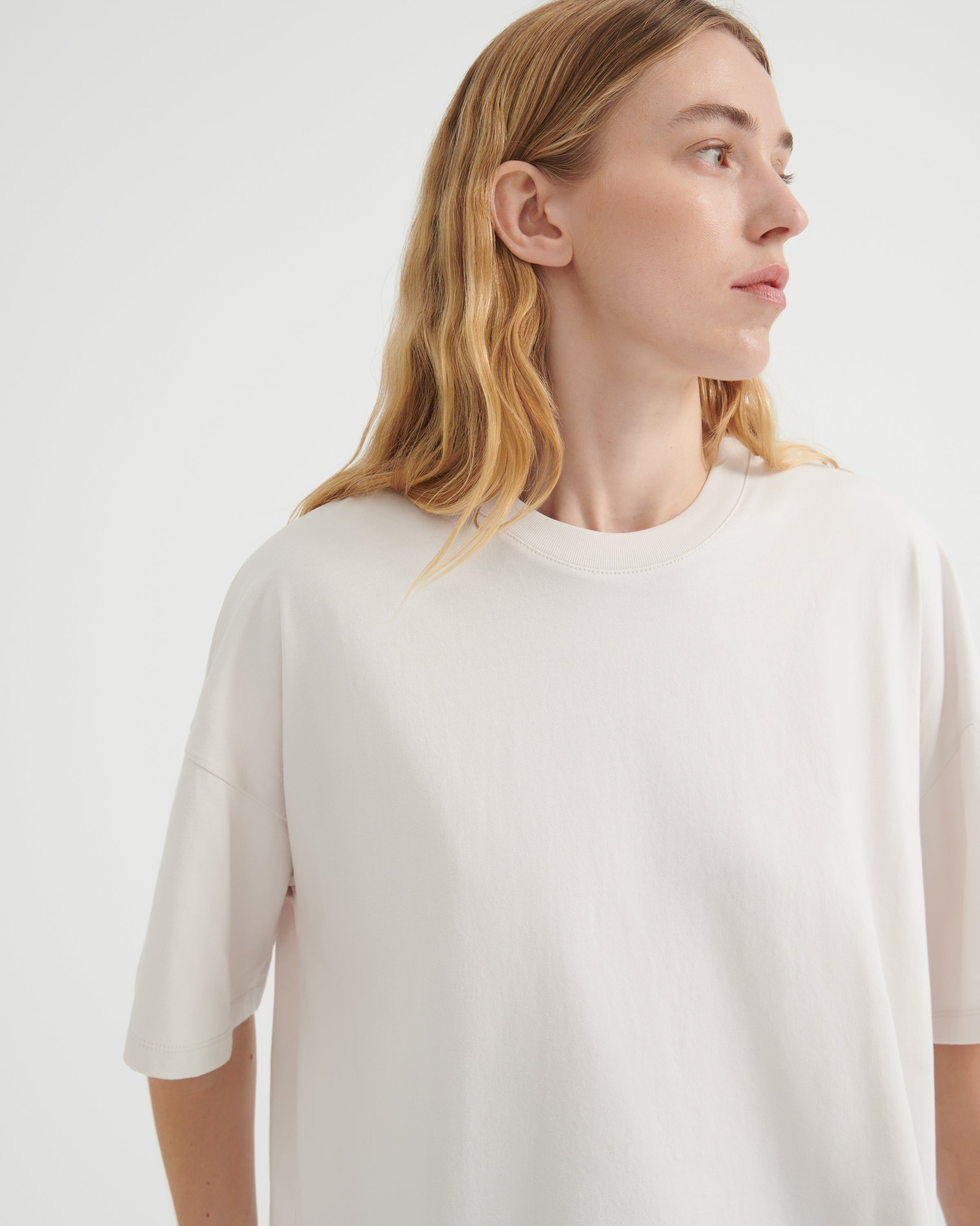 A woman with blonde hair wearing an oversized off-white crewneck t-shirt, shown from the chest up against a plain light gray studio background.