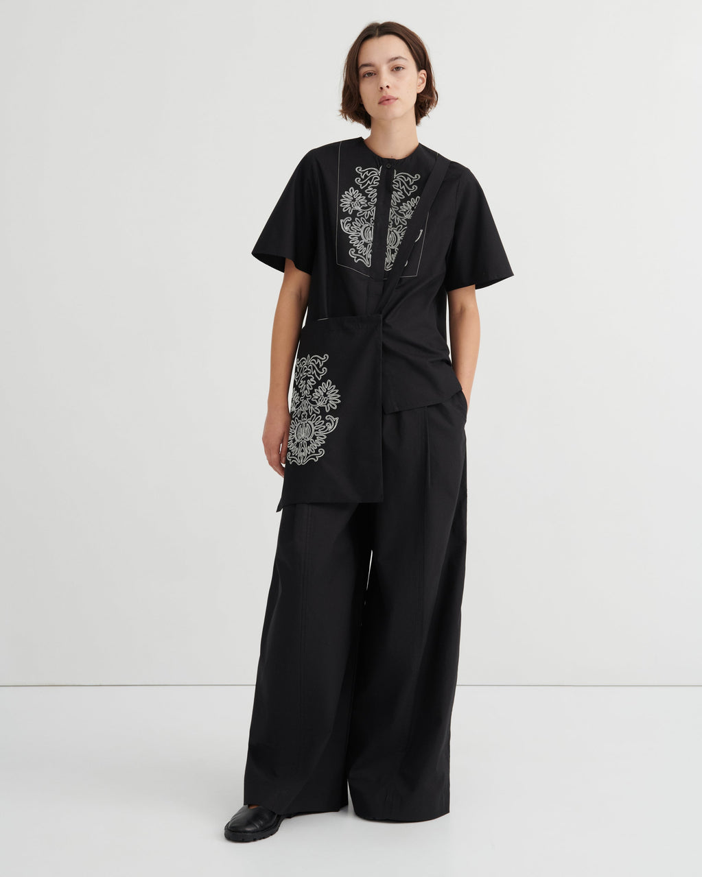 A model stands against a light gray studio background wearing a black short-sleeve top with white floral embroidery and matching black wide-leg pants.