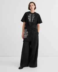 A model stands against a light gray studio background wearing a black short-sleeve top with white floral embroidery and matching black wide-leg pants.