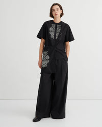 A model stands against a light gray studio background wearing a black short-sleeve top with white floral embroidery and matching black wide-leg pants.