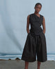 A women wears an asymmetric black top and wide leg pants against a blue background.