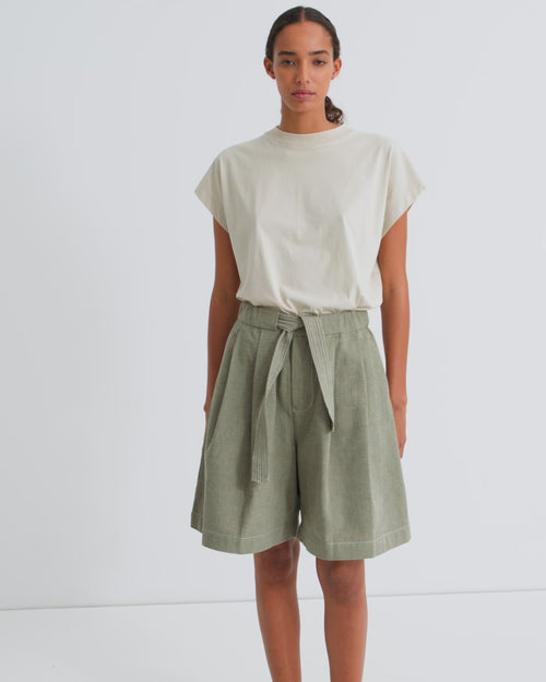 A women wearing a sleeveless off white tee tucked into belted green shorts.