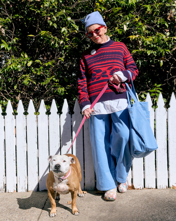 A woman wearing a blue beanie, red and navy patterned sweater, and light blue pants walks a tan dog on a pink leash in front of a white picket fence and green bushes. She is carrying a large light blue tote bag.