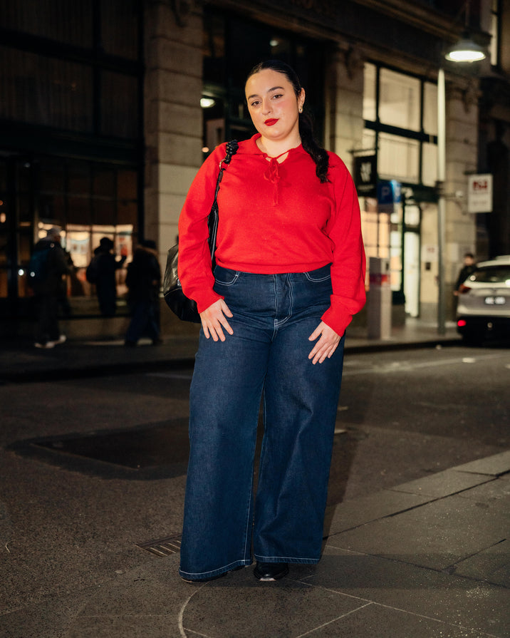 A woman stands on a city street at night wearing a bright red long-sleeve top and dark wide-leg denim jeans, with a black shoulder bag.
