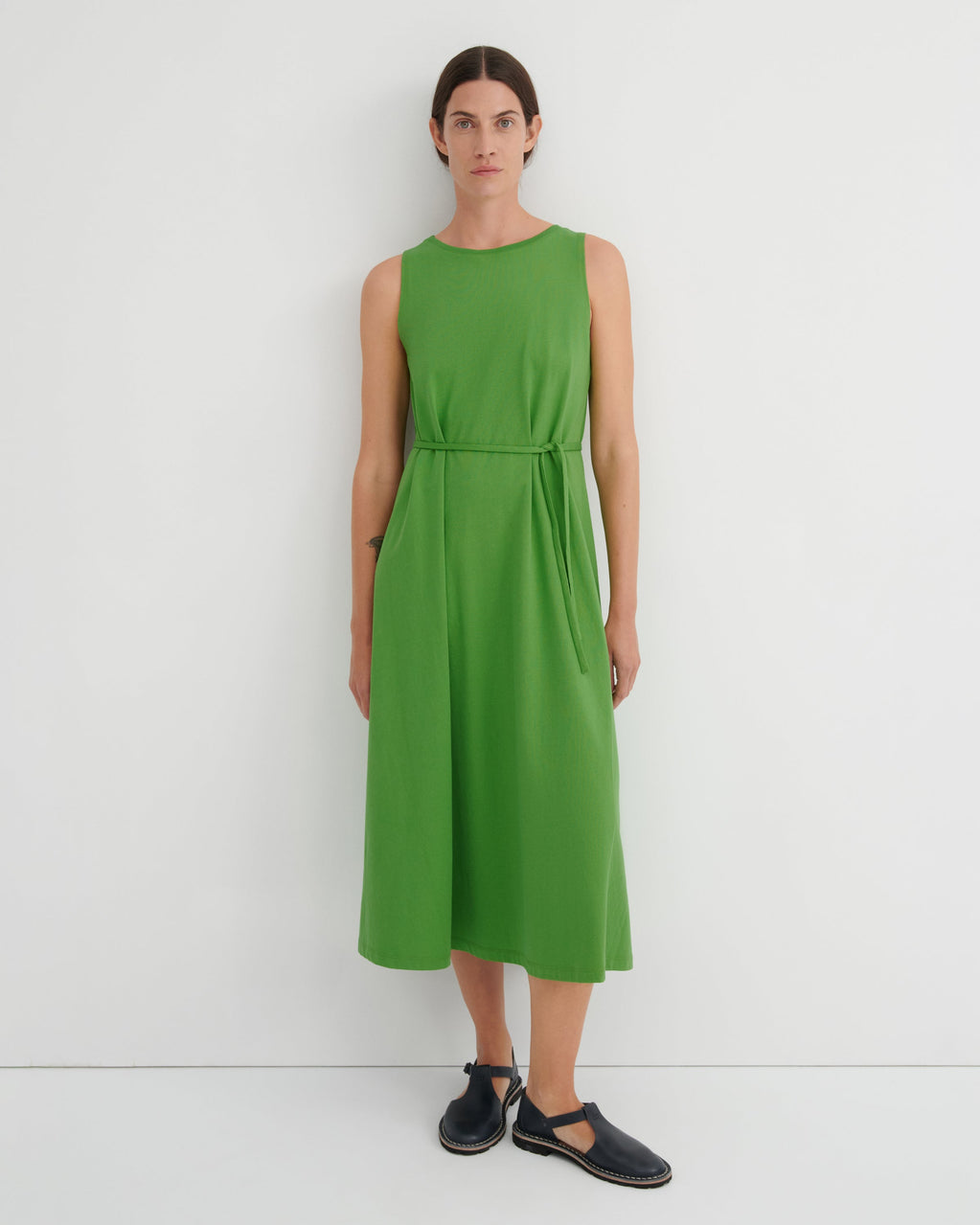 A woman stands against a plain white wall wearing a sleeveless bright green midi dress with a tie waist and dark navy flat shoes.