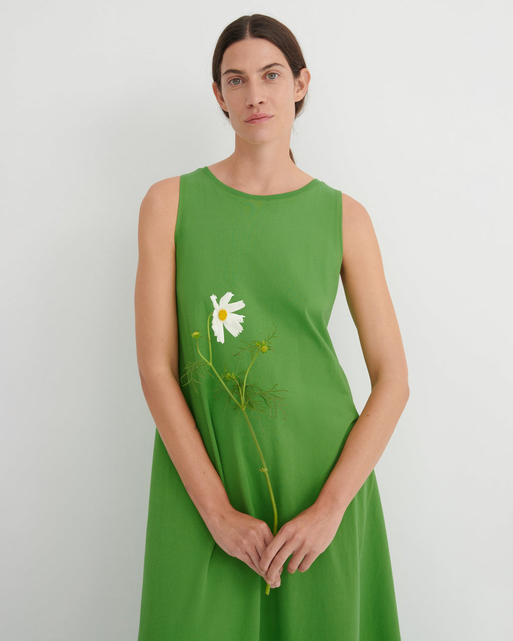 A woman wearing a sleeveless bright green dress with a white daisy graphic on the front, holding a real daisy stem against a plain light gray background.