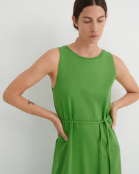 A woman wearing a sleeveless bright green dress with a matching tie belt at the waist, photographed against a plain light gray studio background.