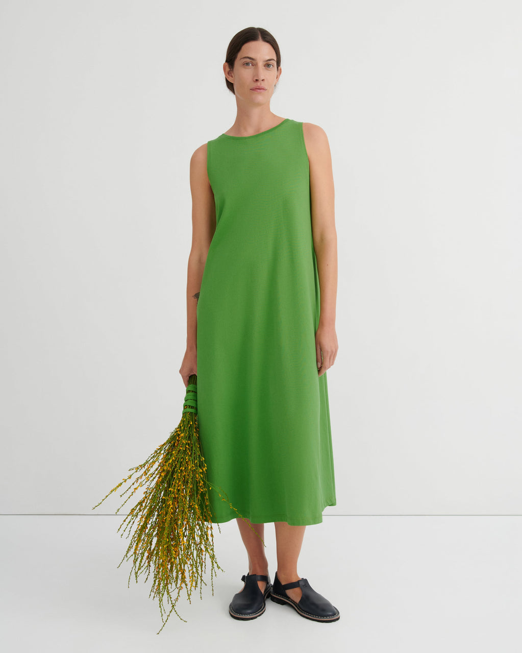 A woman stands against a plain light gray studio background wearing a sleeveless, knee-length bright green dress and dark slip-on shoes, while holding a bunch of yellow-green stems.