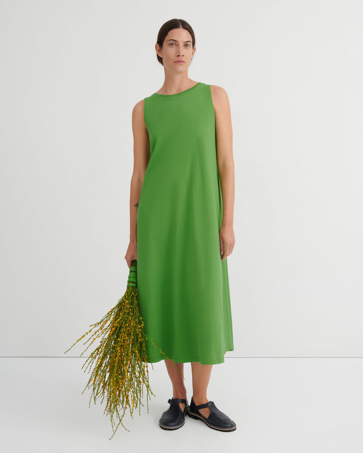 A woman stands against a plain light gray studio background wearing a sleeveless, knee-length bright green dress and dark slip-on shoes, while holding a bunch of yellow-green stems.