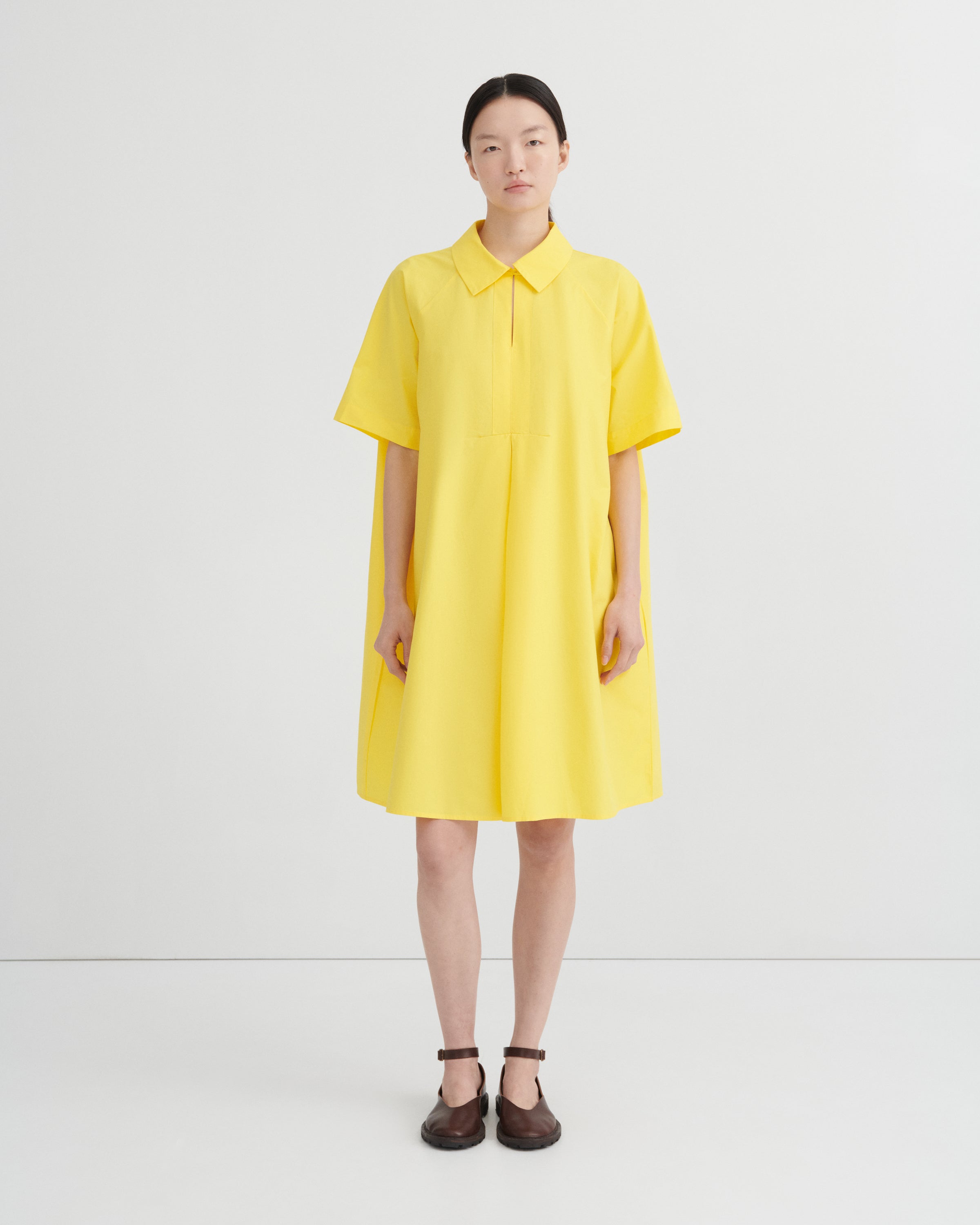 A model stands against a light gray studio background wearing a bright yellow short-sleeve collared dress with a relaxed, oversized fit and dark brown Mary Jane shoes.