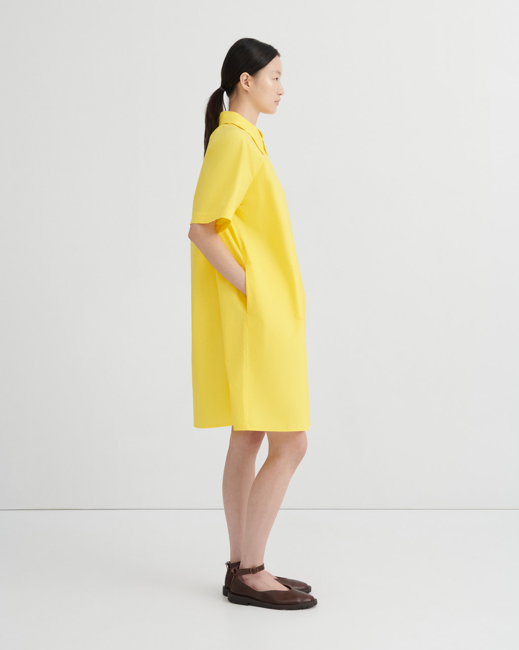 A woman in profile wearing a bright yellow short-sleeve collared dress with side pockets, paired with dark brown flat sandals, against a clean white studio background.