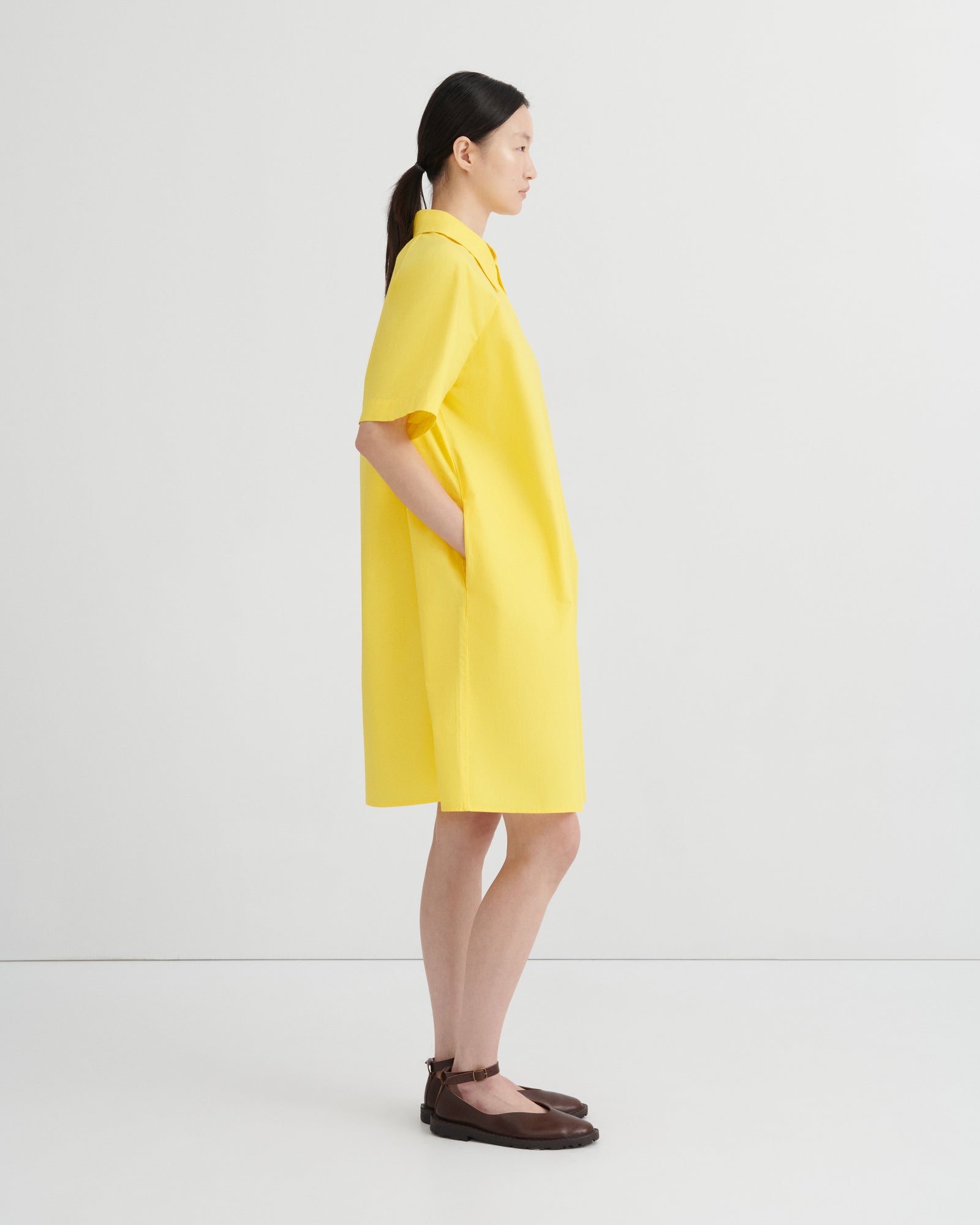 A woman in profile wearing a bright yellow short-sleeve collared dress with side pockets, paired with dark brown flat sandals, against a clean white studio background.