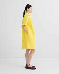 A woman in profile wearing a bright yellow short-sleeve collared dress with side pockets, paired with dark brown flat sandals, against a clean white studio background.