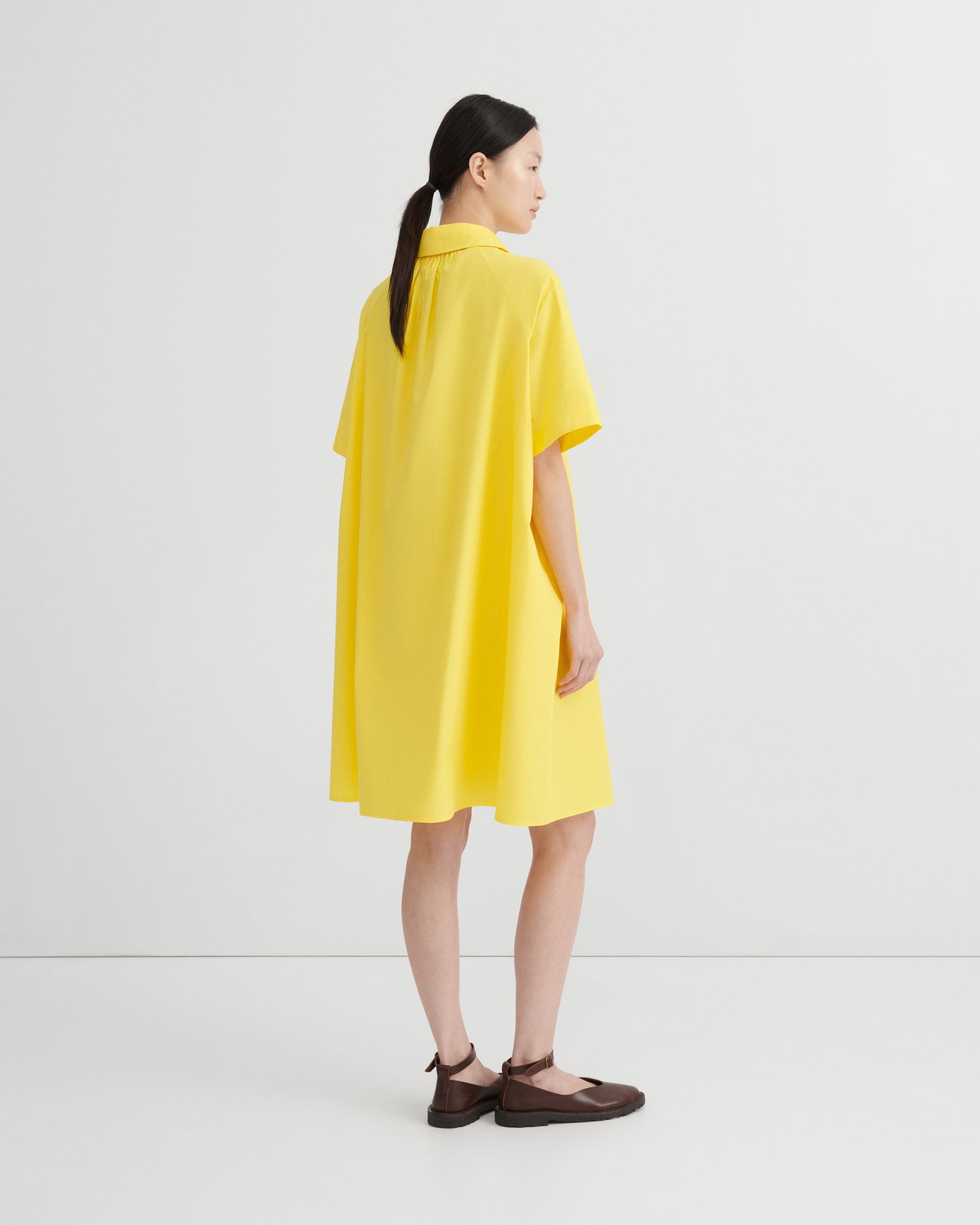 A woman wearing a bright yellow short-sleeve collared dress, shown from the back in a studio setting with a light gray background.