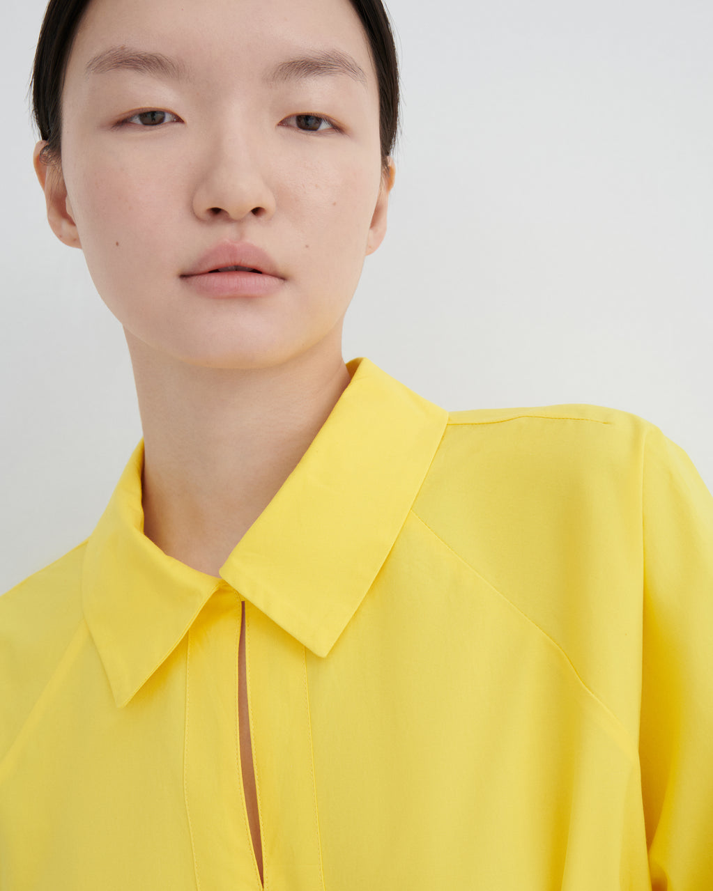 A close-up of a model wearing a bright yellow collared blouse against a plain light gray background.