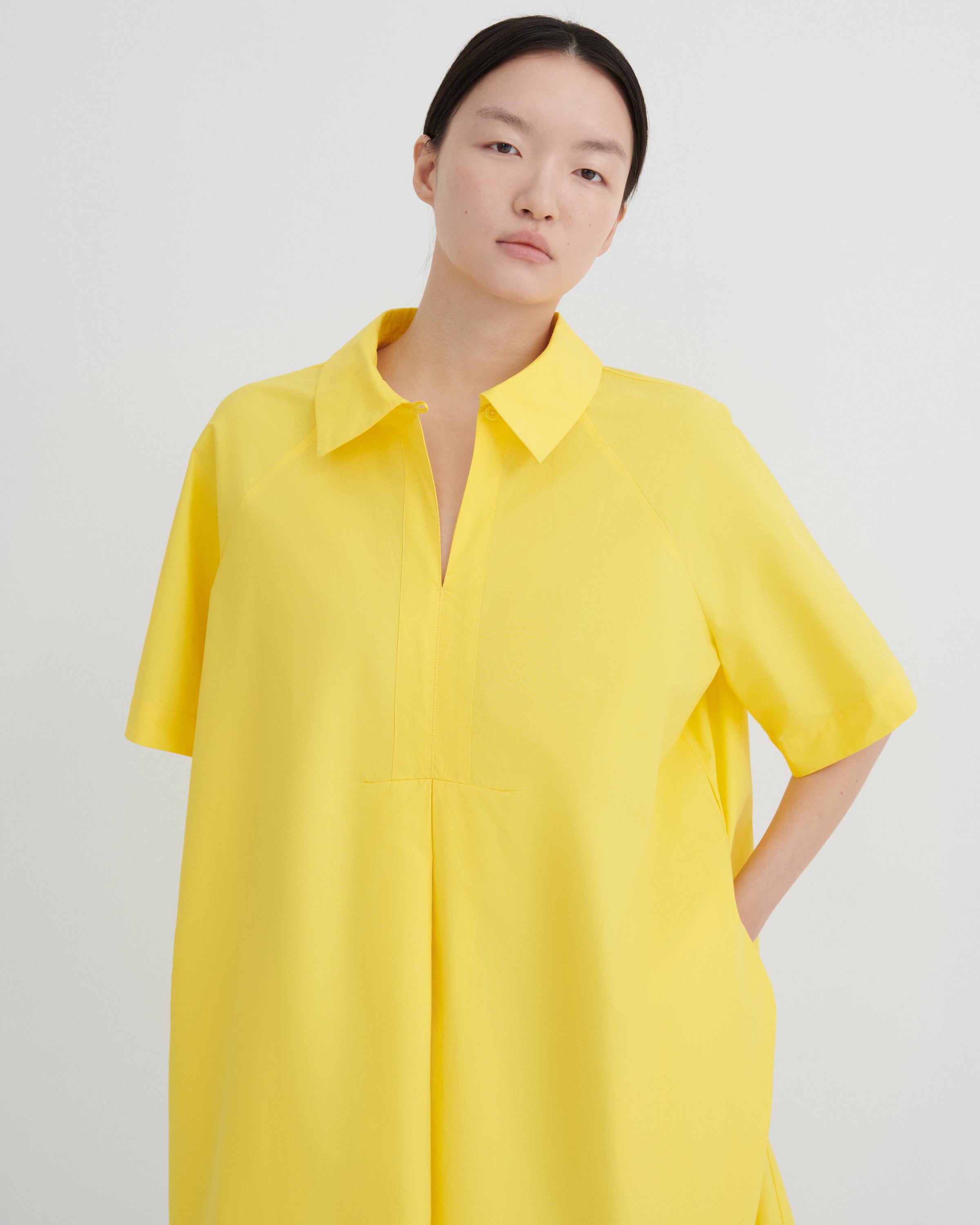 A woman wearing a bright yellow short-sleeve collared shirt dress, photographed against a clean light gray studio background.