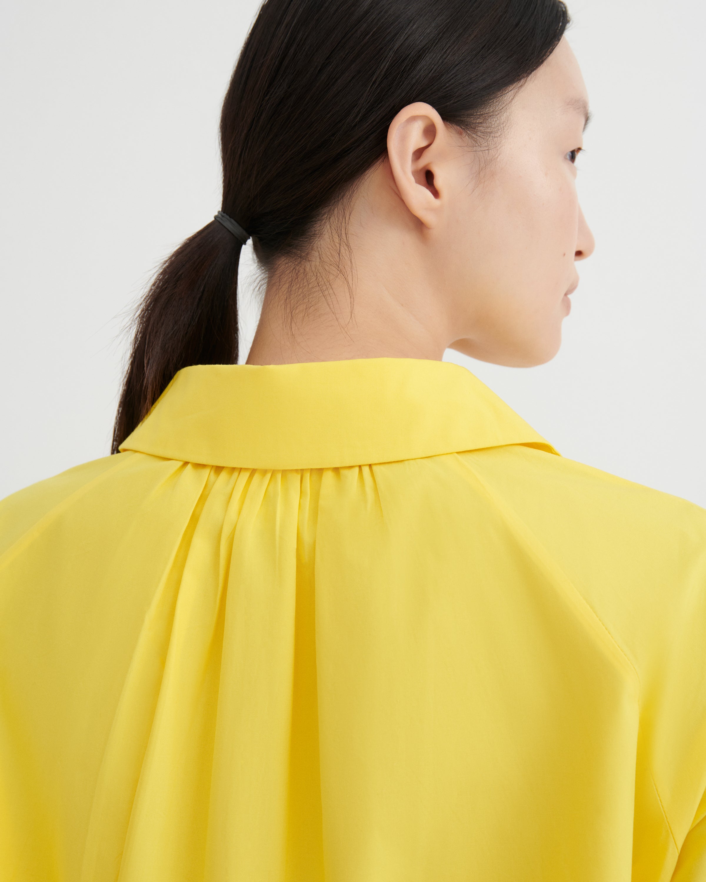 Close-up side profile of a model wearing a bright yellow blouse with a wide collar and gathered neckline, against a plain light gray background.