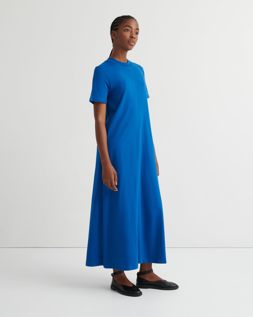 A model wearing a bright blue short-sleeve midi dress with a crew neck, paired with black flat shoes, standing against a plain white studio background.