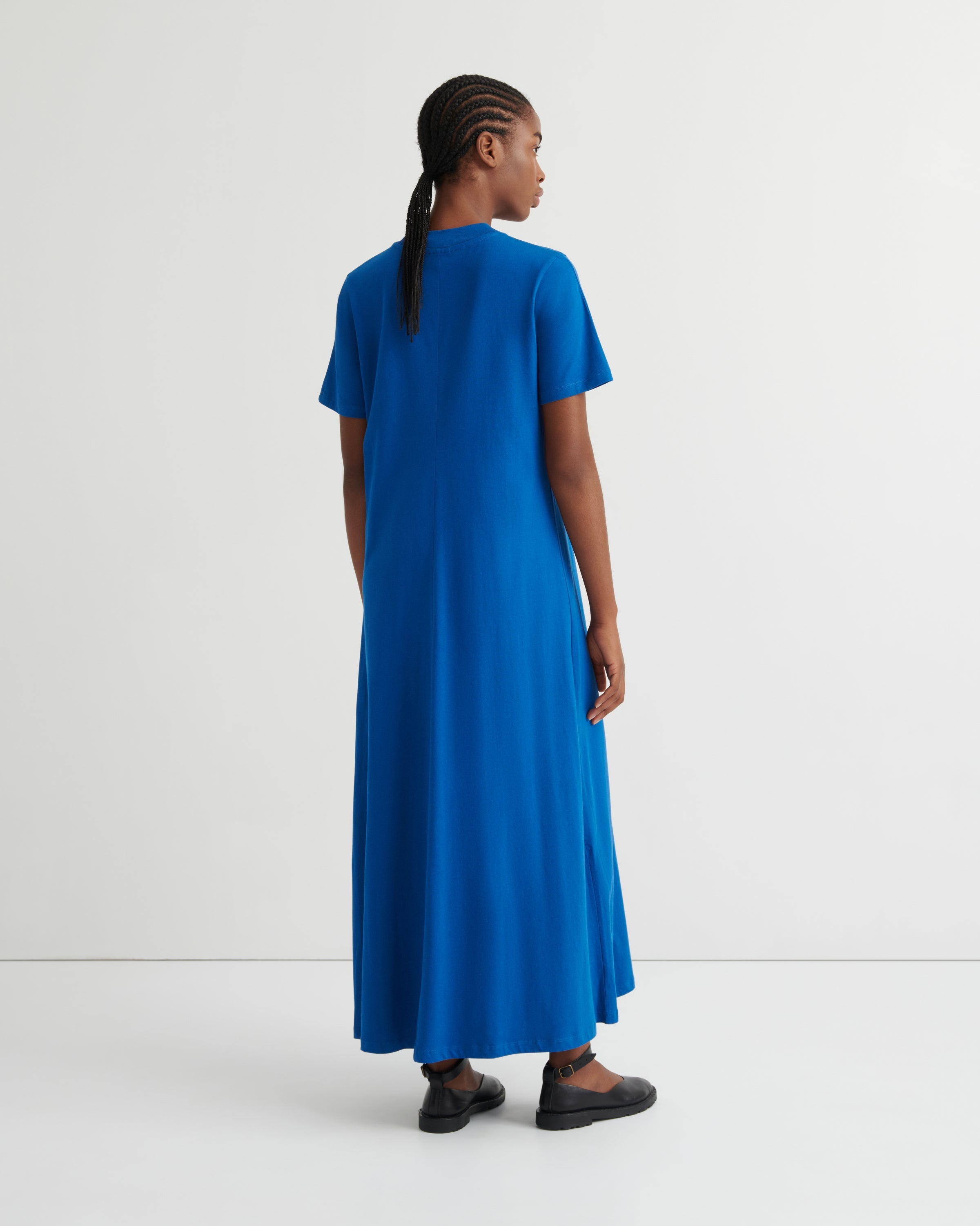A woman wearing a bright blue short-sleeve maxi dress, shown from the back in a studio setting with a white background.