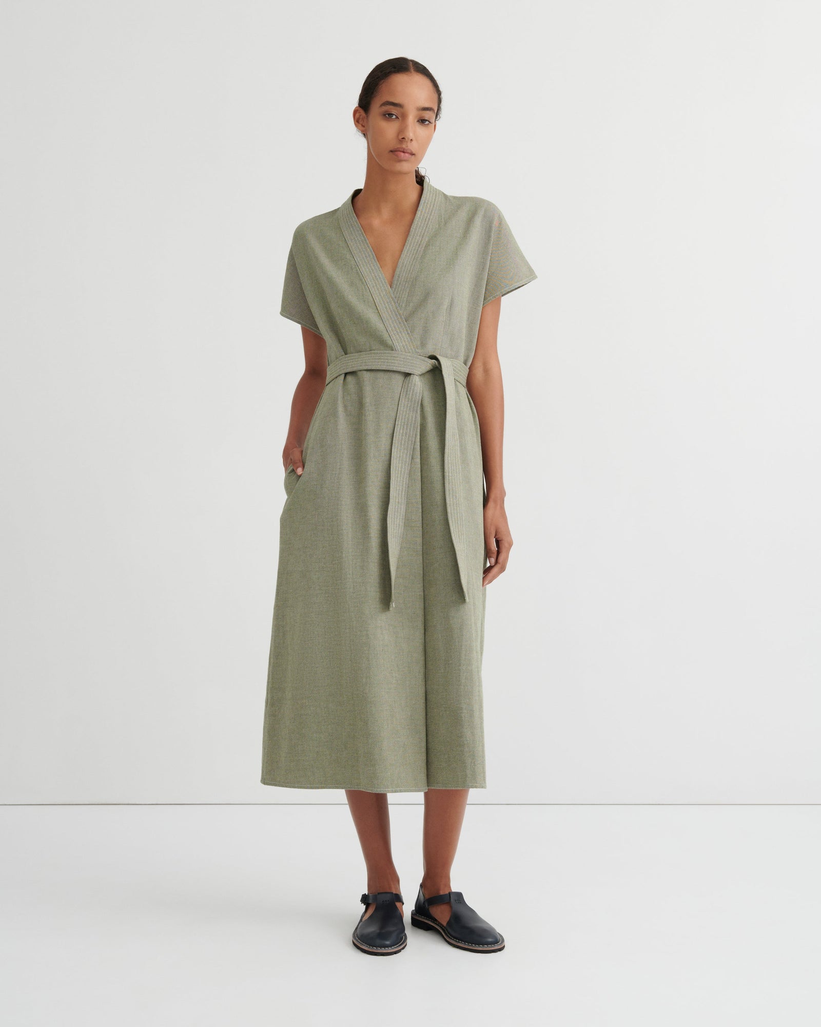 A woman modeling a sage green short-sleeve wrap dress with a matching waist tie, standing against a plain white studio background.