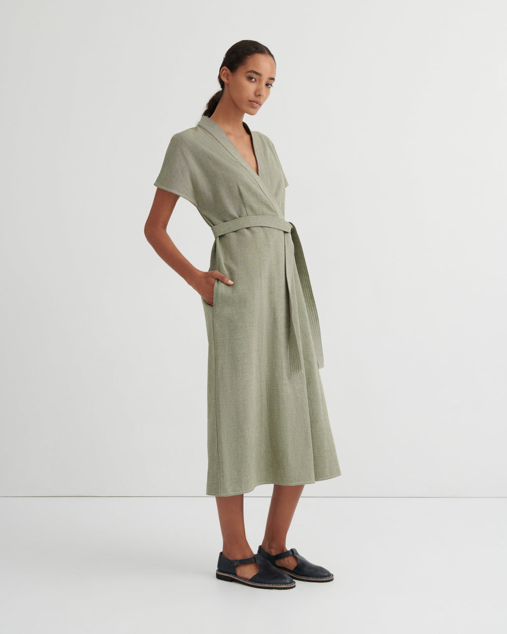 A woman modeling a sage green linen wrap dress with short sleeves, a V-neckline, a matching waist tie, and side pockets, paired with dark navy Mary Jane flats, against a plain white studio background.