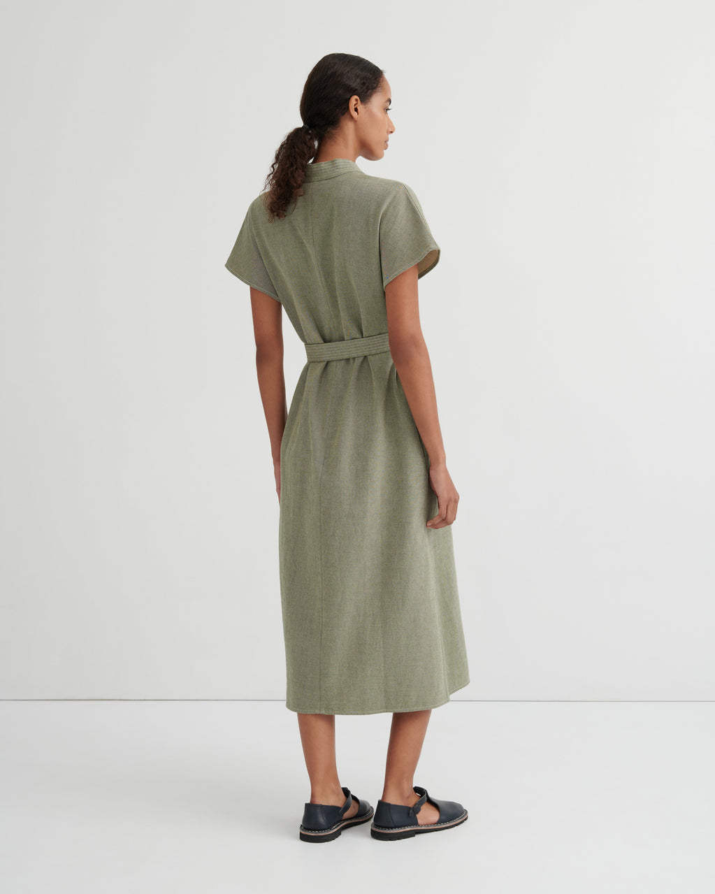 A woman shown from the back wearing a short-sleeved olive green midi dress with a matching waist tie, paired with dark sandals, against a plain light gray studio background.