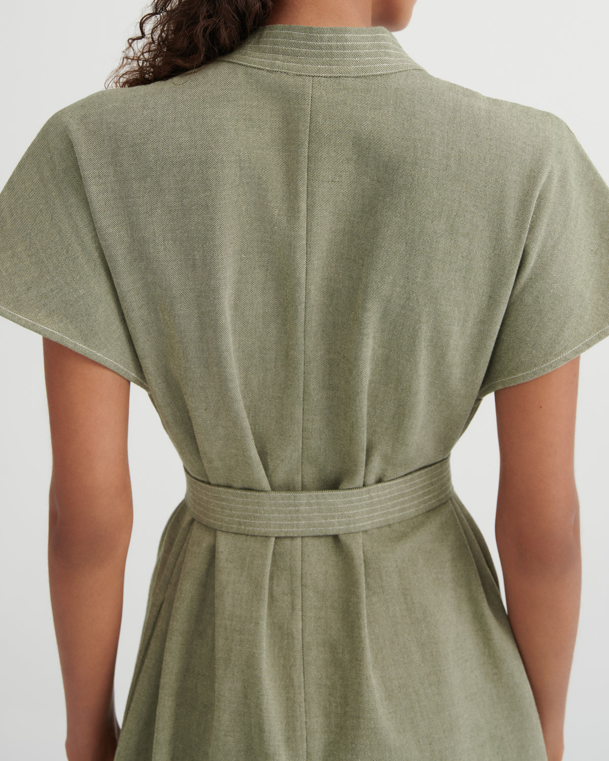 Back view of a woman wearing a short-sleeve sage green dress with a matching waist tie belt, shown against a plain light gray background.