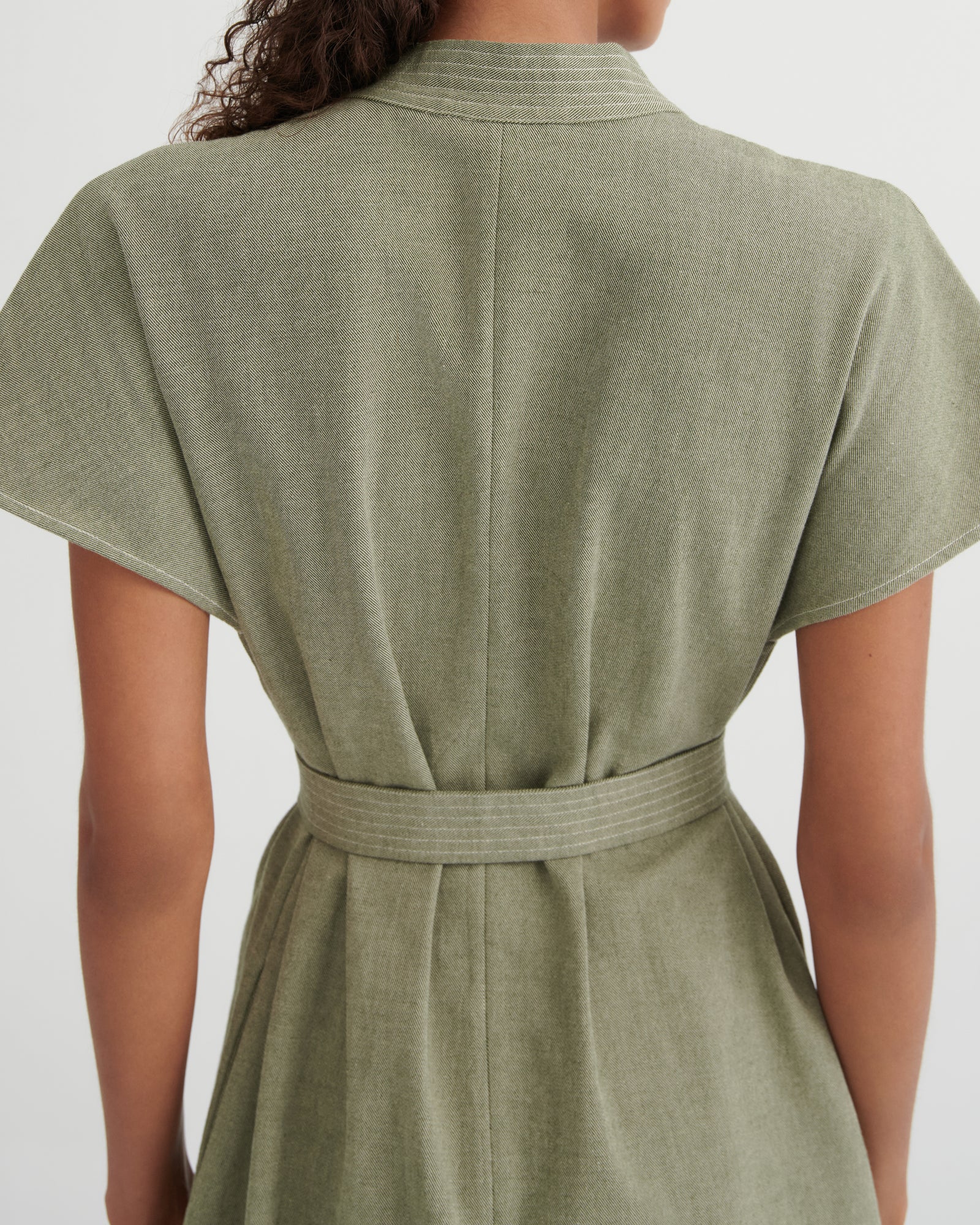 Back view of a woman wearing a short-sleeve sage green dress with a matching waist tie belt, shown against a plain light gray background.