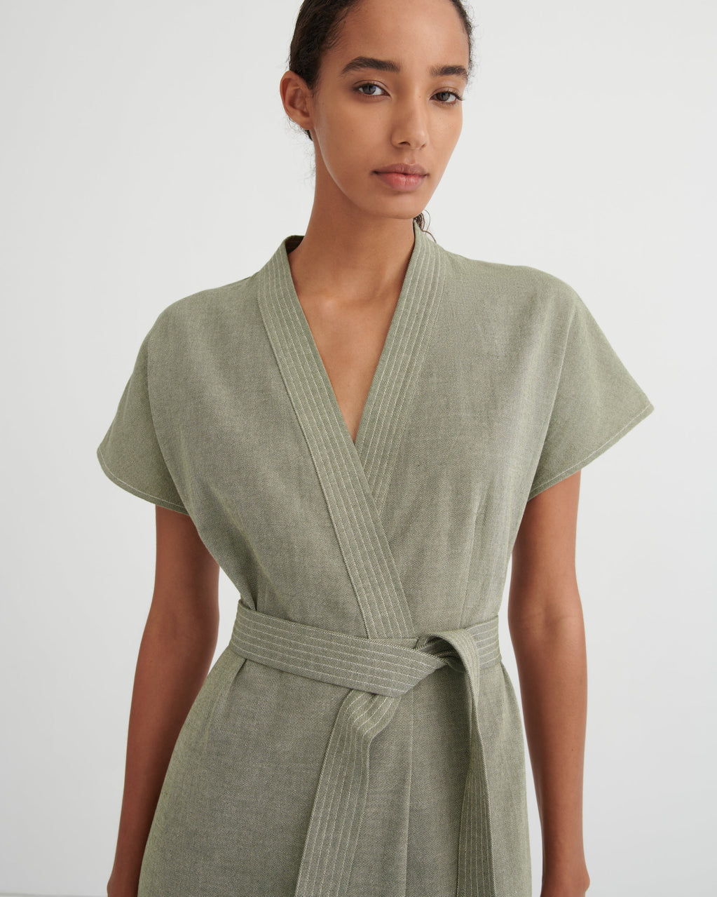 A woman wearing a sage green short-sleeve wrap dress with a matching tie belt, photographed against a plain light gray studio background.