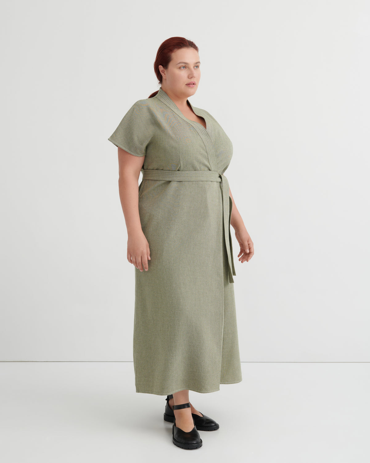 A woman modeling a sage green wrap midi dress with short sleeves and a matching waist tie, paired with black Mary Jane shoes, against a plain light gray studio background.