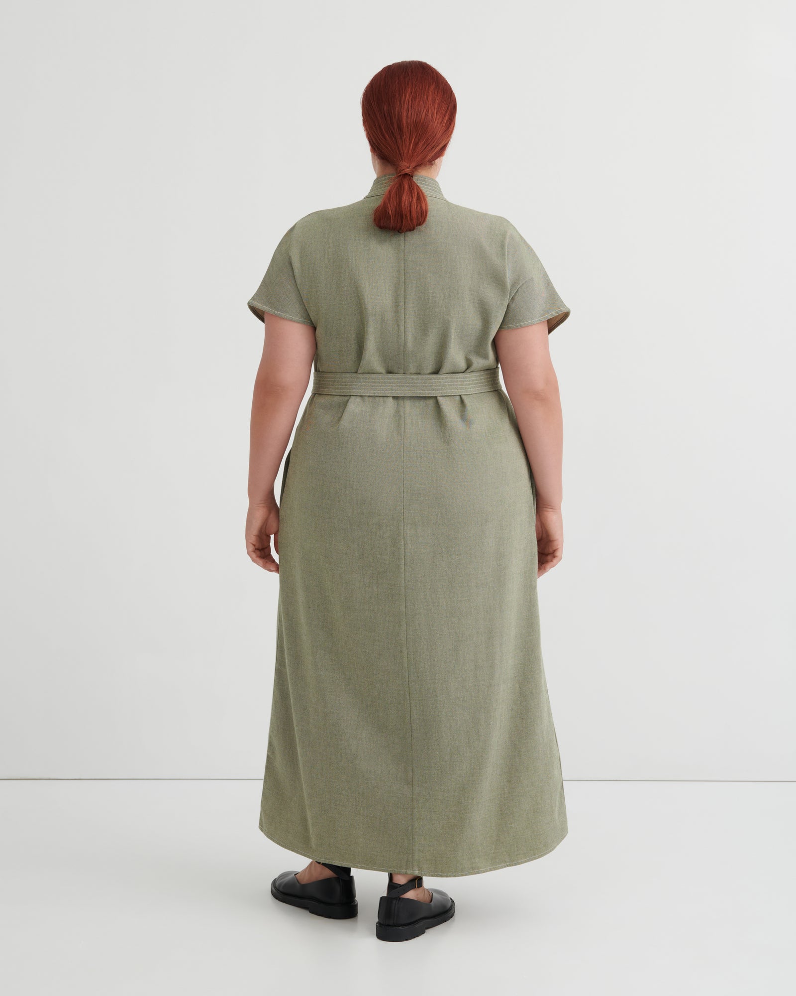A person shown from the back wearing a short-sleeve, knee-length olive green dress with a matching waist tie, photographed against a clean light gray studio background.