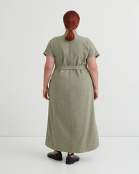 A person shown from the back wearing a short-sleeve, knee-length olive green dress with a matching waist tie, photographed against a clean light gray studio background.
