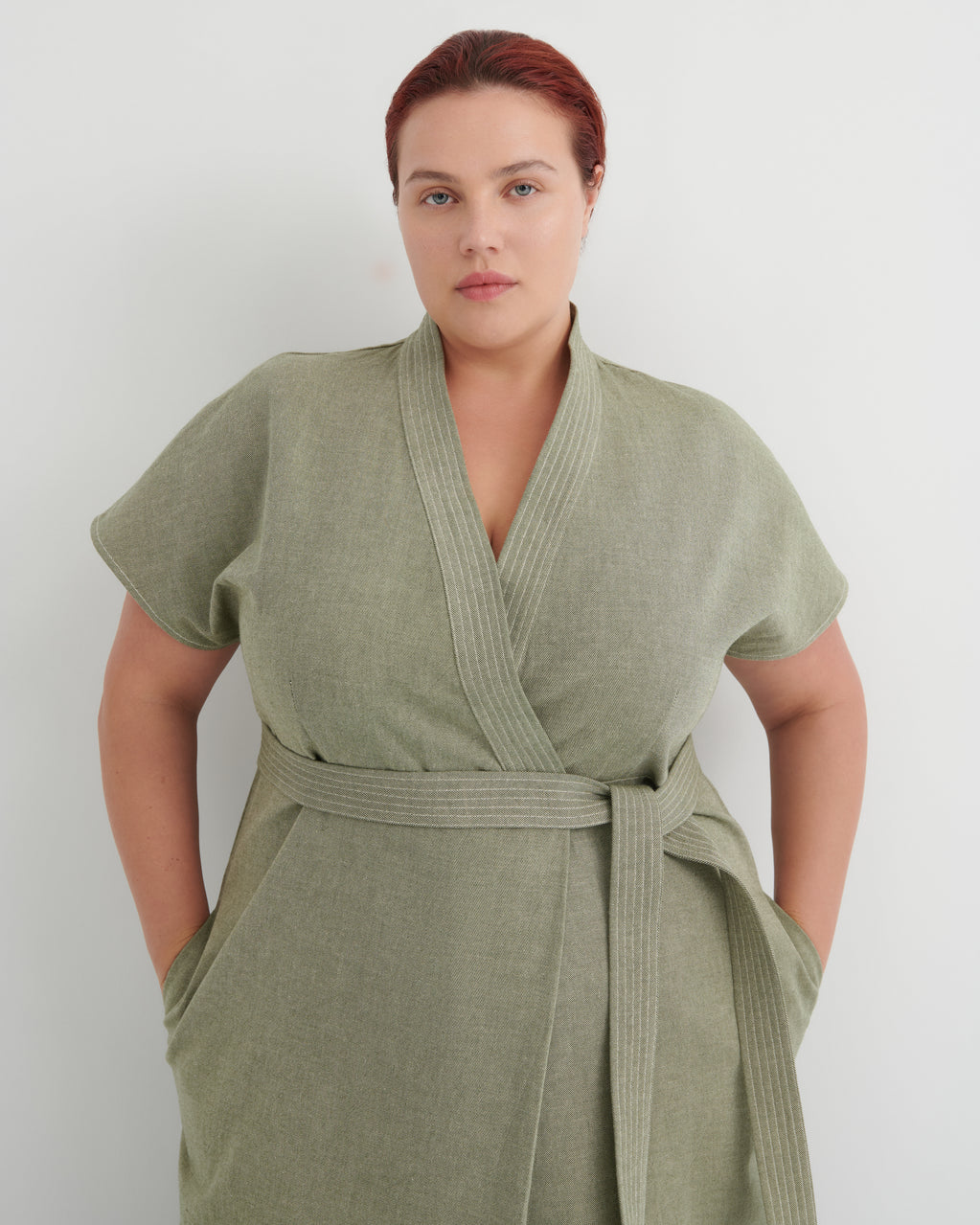 A woman wearing a sage green short-sleeve wrap robe with a matching waist tie, standing against a plain light gray background.