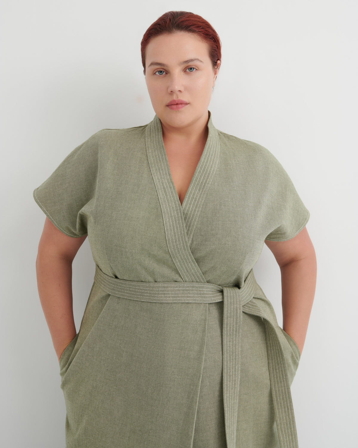 A woman wearing a sage green short-sleeve wrap robe with a matching waist tie, standing against a plain light gray background.