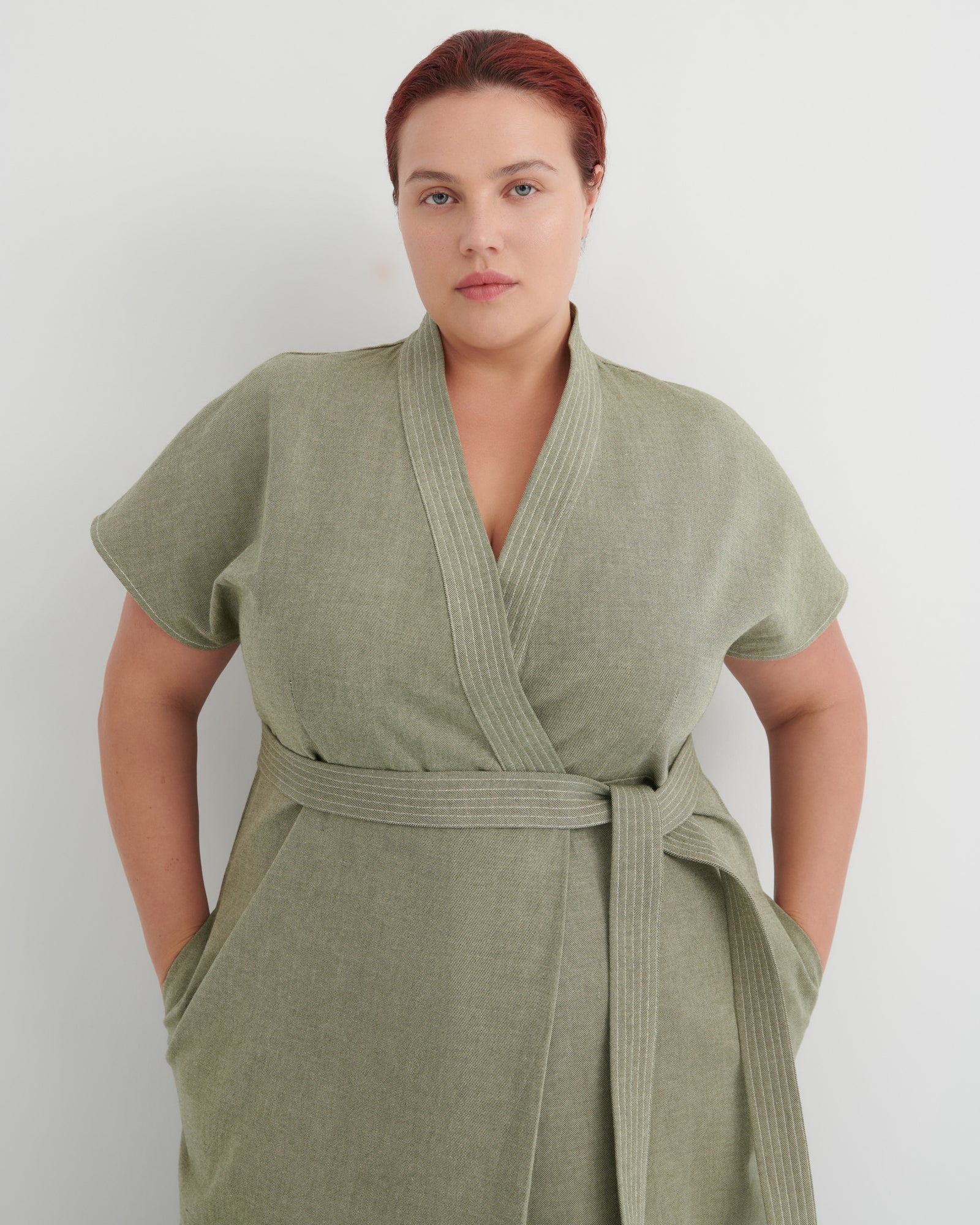 A woman wearing a sage green short-sleeve wrap robe with a matching waist tie, standing against a plain light gray background.
