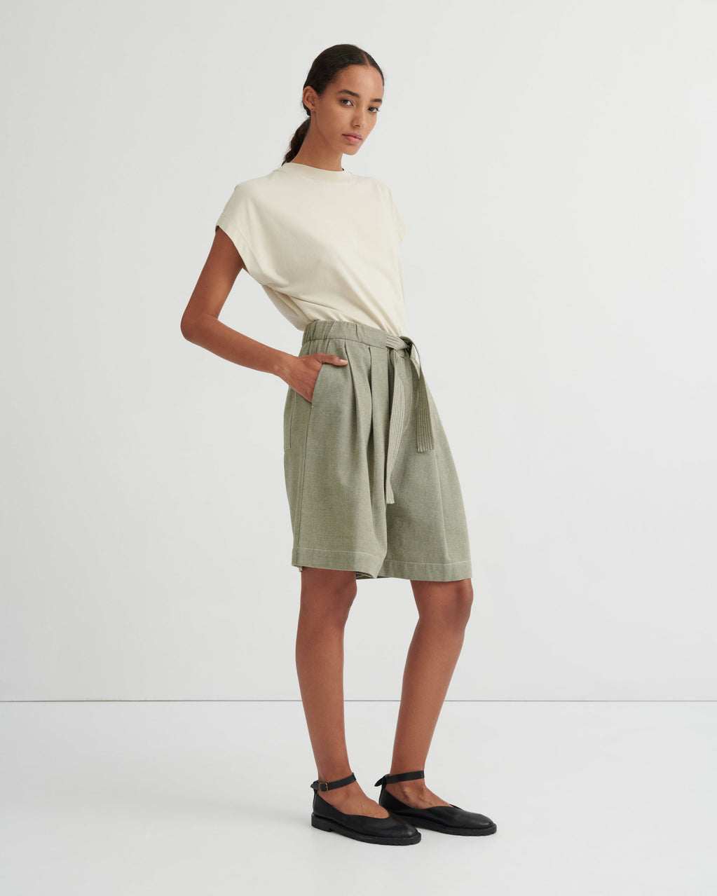 A model stands against a clean white studio background wearing a cream-colored short-sleeve t-shirt and sage green linen shorts with a matching tie belt, paired with black flat shoes.