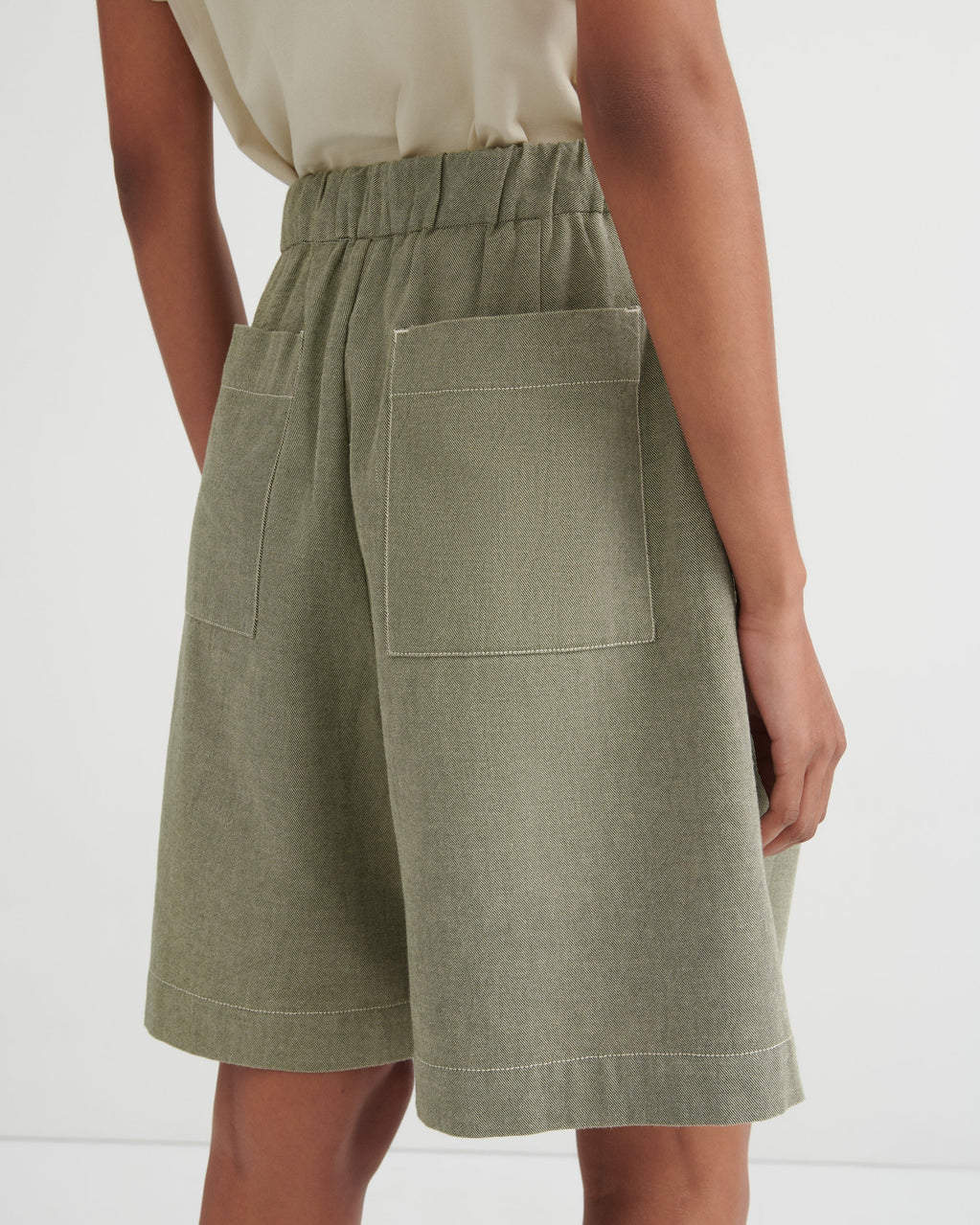 A close-up back view of a model wearing sage green linen shorts with an elastic waistband and large patch pockets, paired with a light beige top against a white studio background.