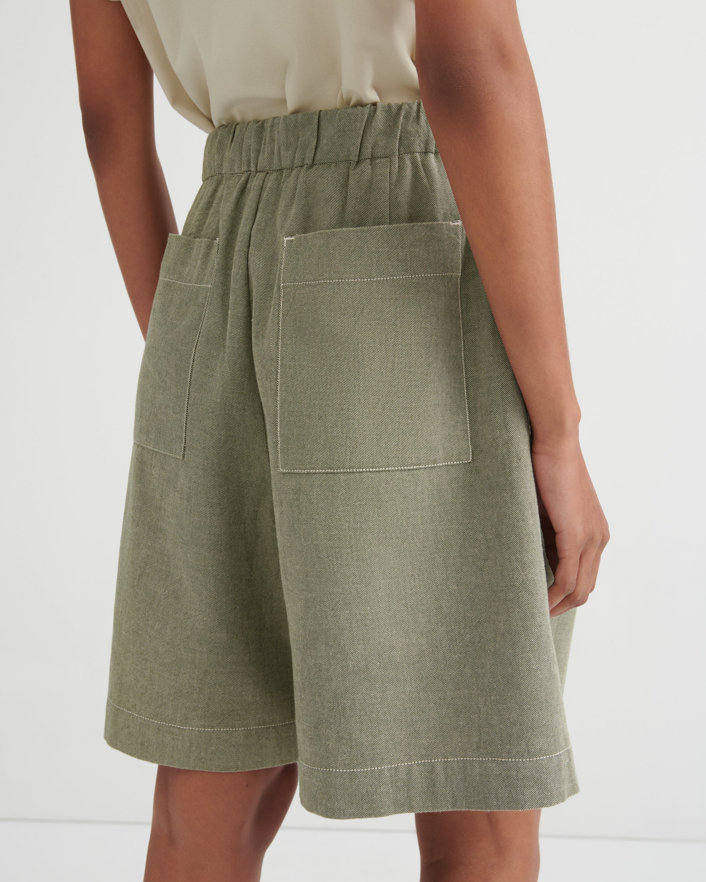 A close-up back view of a model wearing sage green linen shorts with an elastic waistband and large patch pockets, paired with a light beige top against a white studio background.