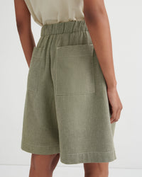 A close-up back view of a model wearing sage green linen shorts with an elastic waistband and large patch pockets, paired with a light beige top against a white studio background.