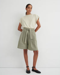 A model stands against a plain white studio wall wearing a cream short-sleeve top and light sage green high-waisted linen shorts with a matching waist tie, paired with black Mary Jane shoes.
