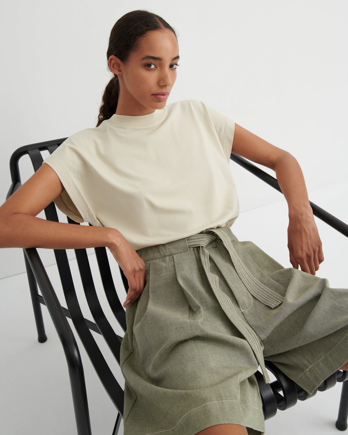A woman seated on a black metal chair wearing a cream short-sleeve mock-neck top and olive green high-waisted wide-leg shorts with a tie belt, against a clean white studio background.